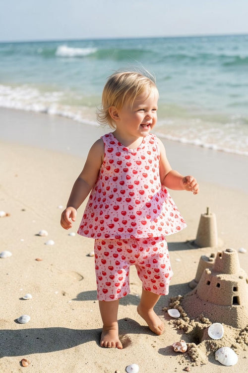 Child in a heart-patterned outfit standing on a sandy beach with ocean waves in the background.